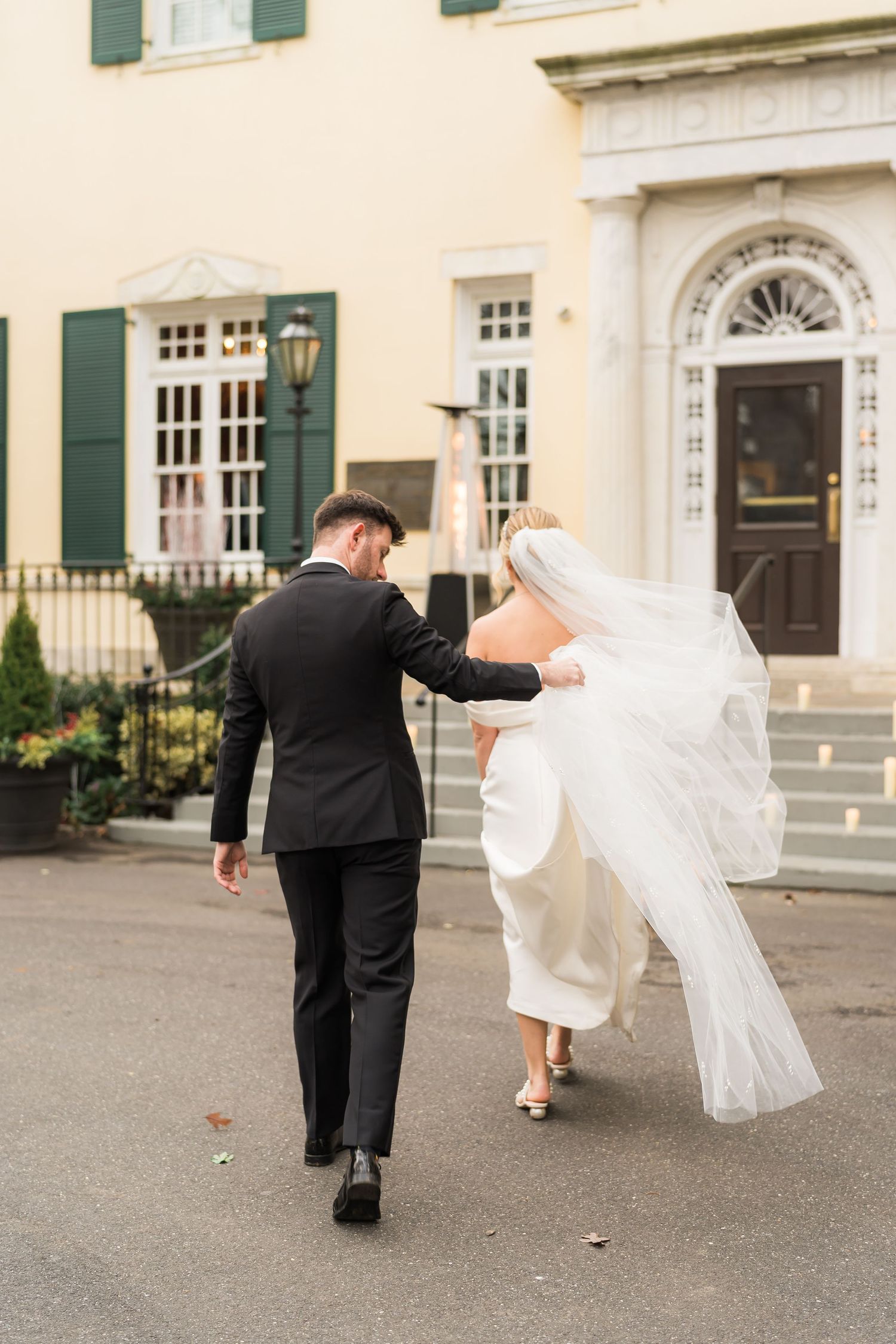 A couple walking outside a historic mansion with green shutters as the bride's veil flows in the breeze.