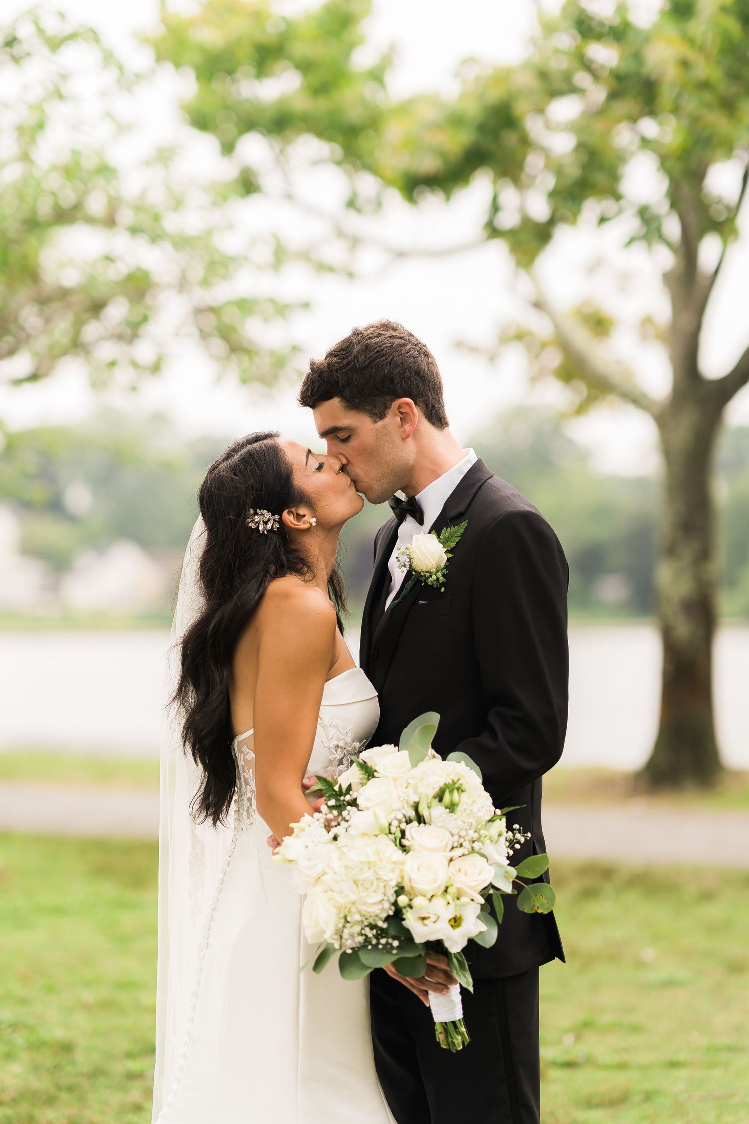 A romantic wedding portrait shows a couple kissing while holding a white floral bouquet outdoors.