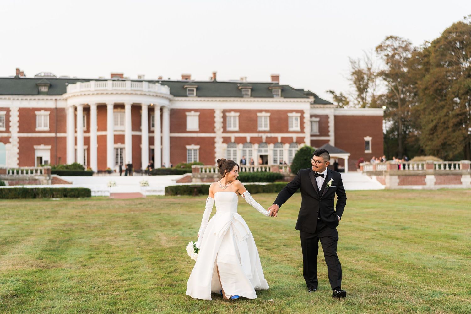 A couple walks across the lawn of a grand colonial style mansion.