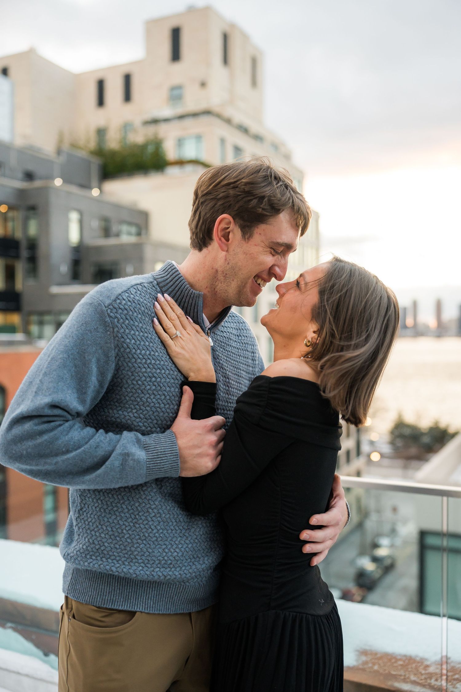 Joyful couple shares intimate moment on rooftop balcony with city buildings in background at sunset.