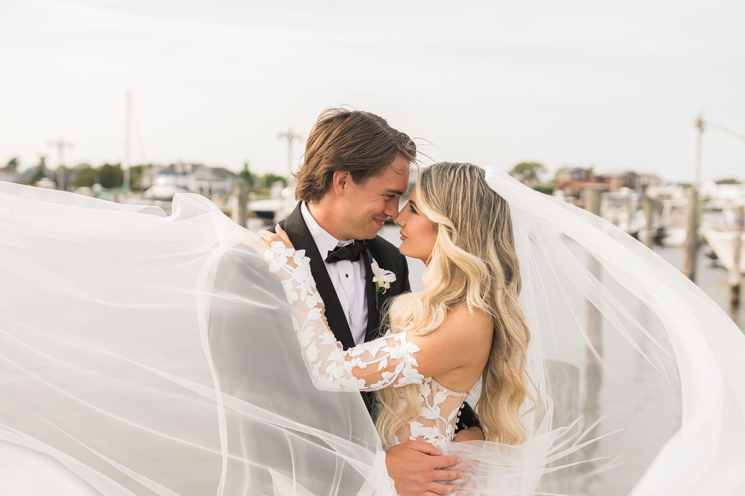 A romantic wedding couple embraces as the bride's veil flows in the wind at a waterfront marina.
