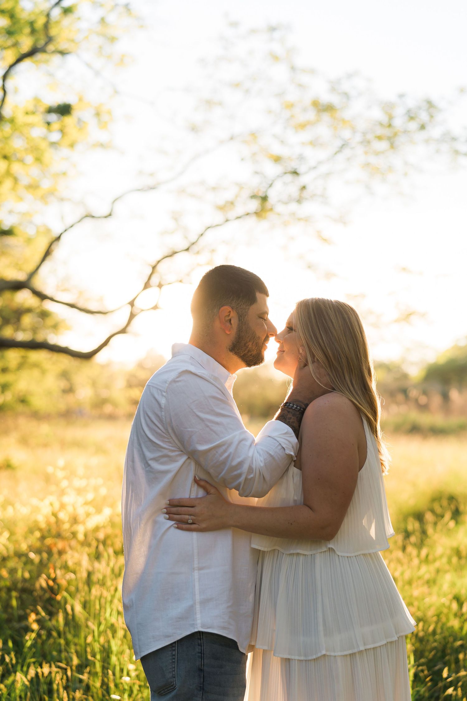 Romantic couple embraces in a sunlit meadow at golden hour with trees in the background.