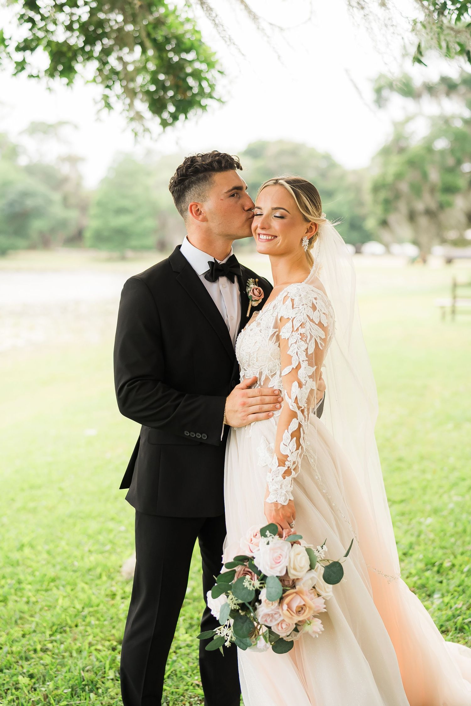 Romantic outdoor wedding portrait on a sunny day with lace dress, black tuxedo, and blush floral bouquet.