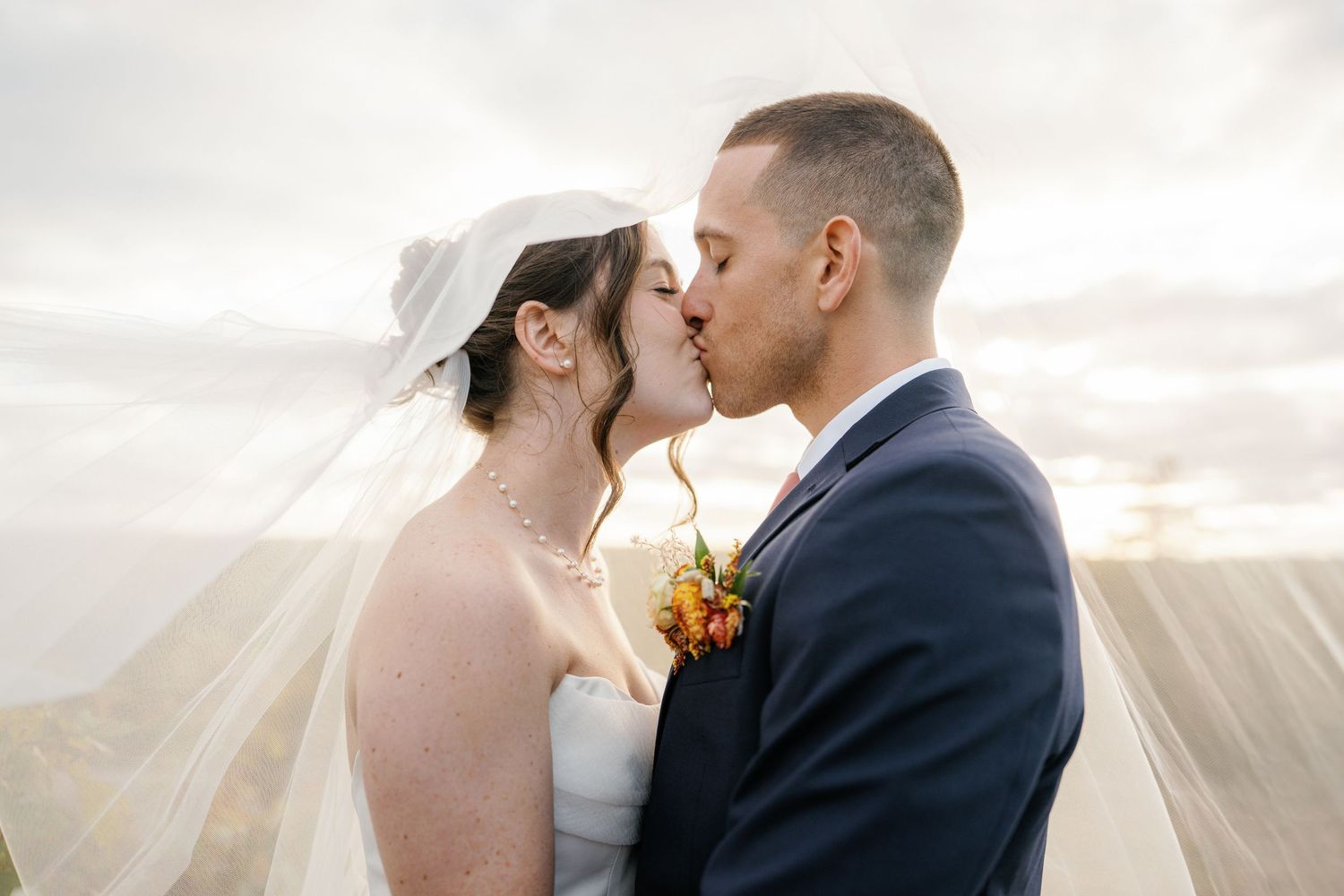 A romantic wedding couple embraces in front of a glowing sunset while a sheer veil floats in the breeze.