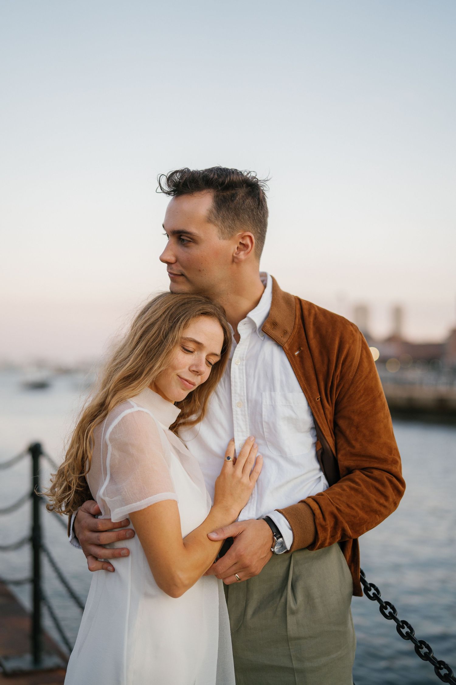 A romantic couple embraces at sunset by the waterfront, with boats and a coastal view in the background.