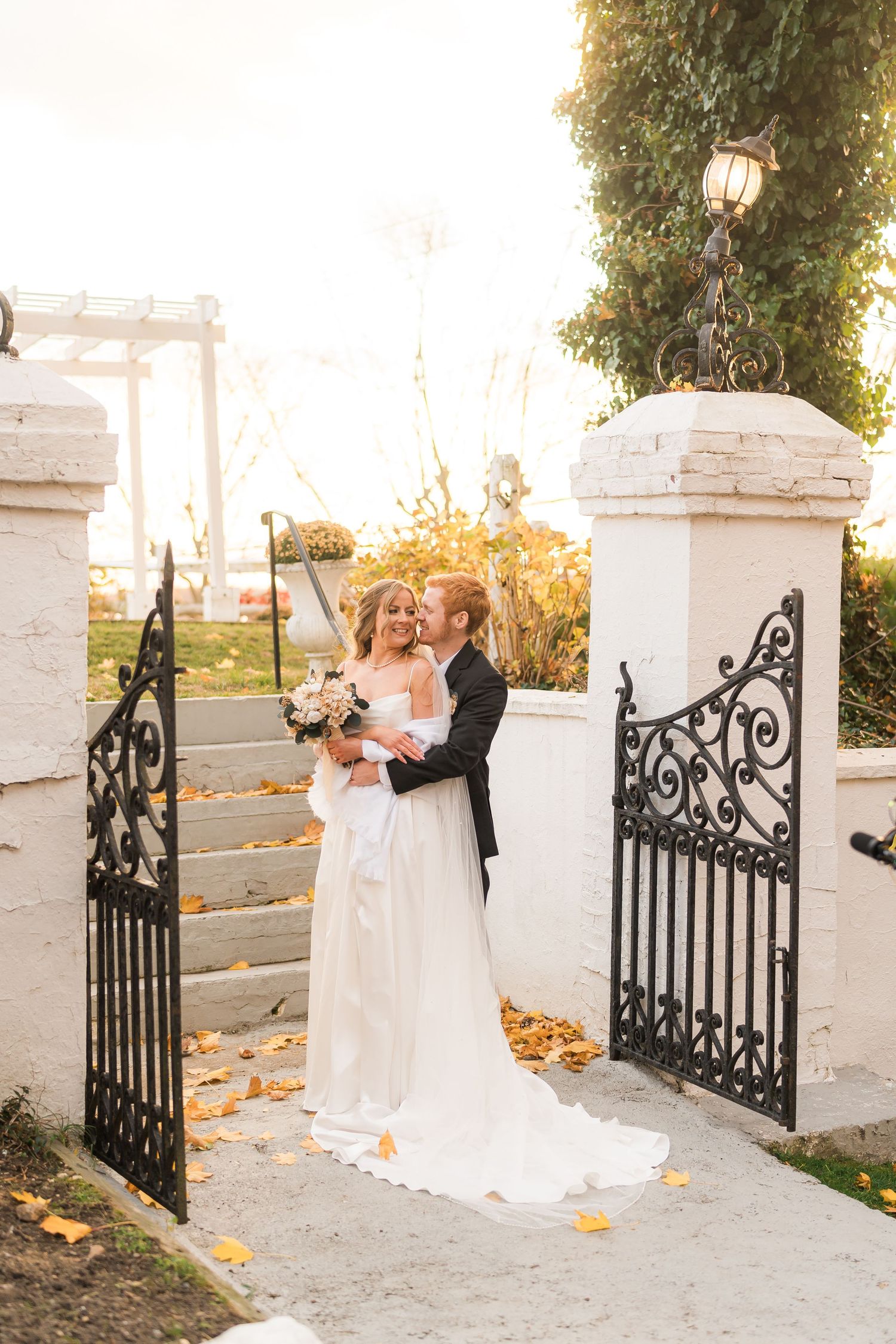 A couple poses between elegant white pillars and ornate black iron gates on their wedding day as autumn leaves scatter around them.