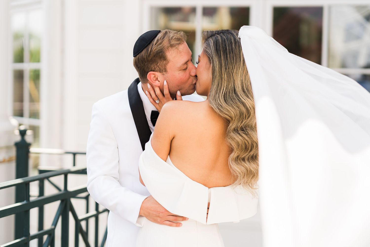 A romantic wedding couple shares a kiss outdoors, with the bride wearing a white strapless gown and flowing veil.