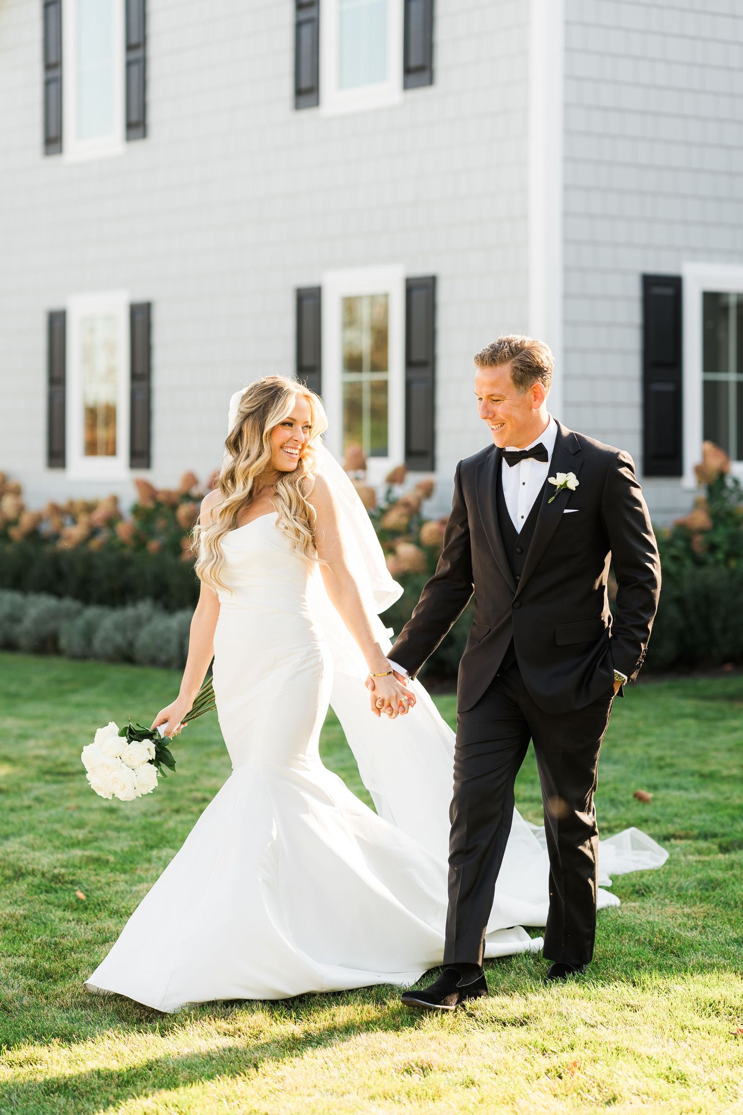 Newlywed couple walking on grass outside white house with bride's dress flowing in sunlight.