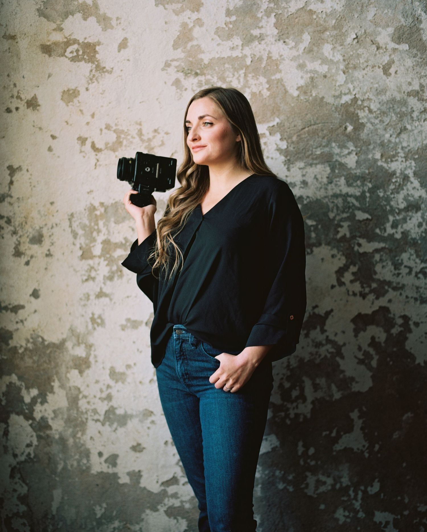 A photographer in black top and blue jeans poses with a vintage camera against a weathered concrete wall.