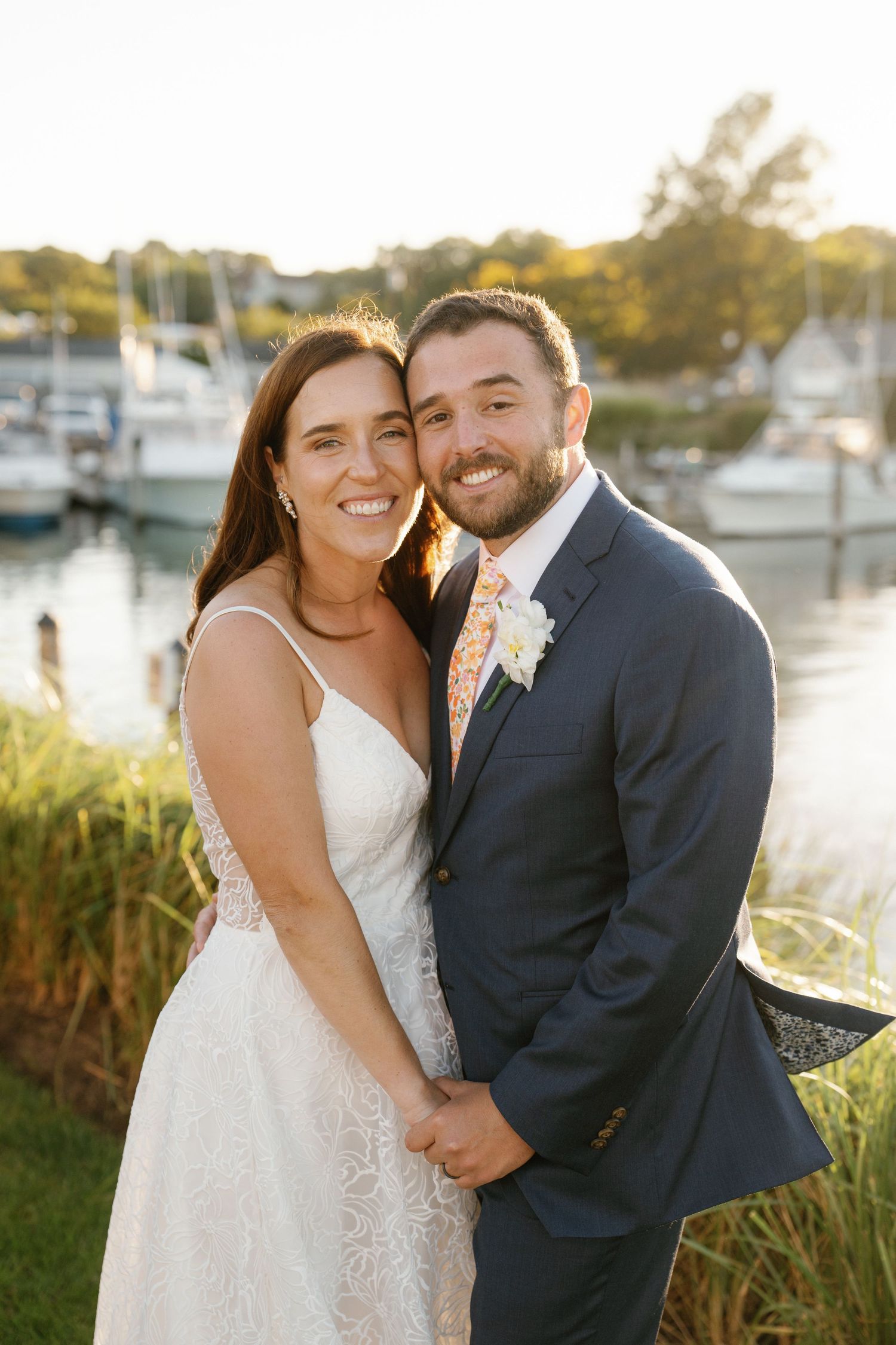 A couple in wedding attire smile together at sunset by a marina with boats and golden light in the background.