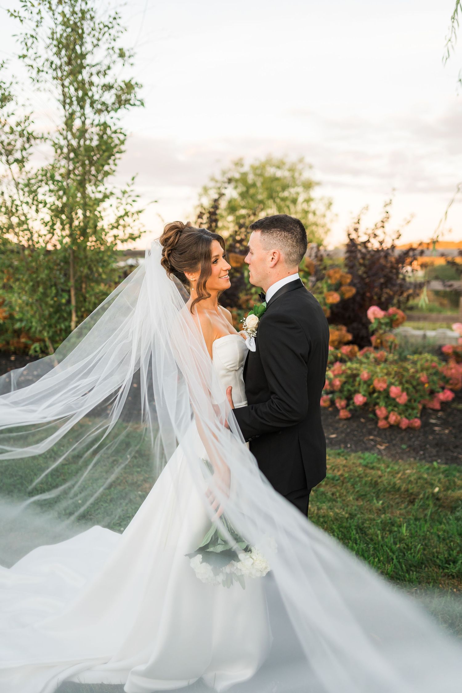 A couple stands close together at sunset with a long flowing white veil and floral garden backdrop.