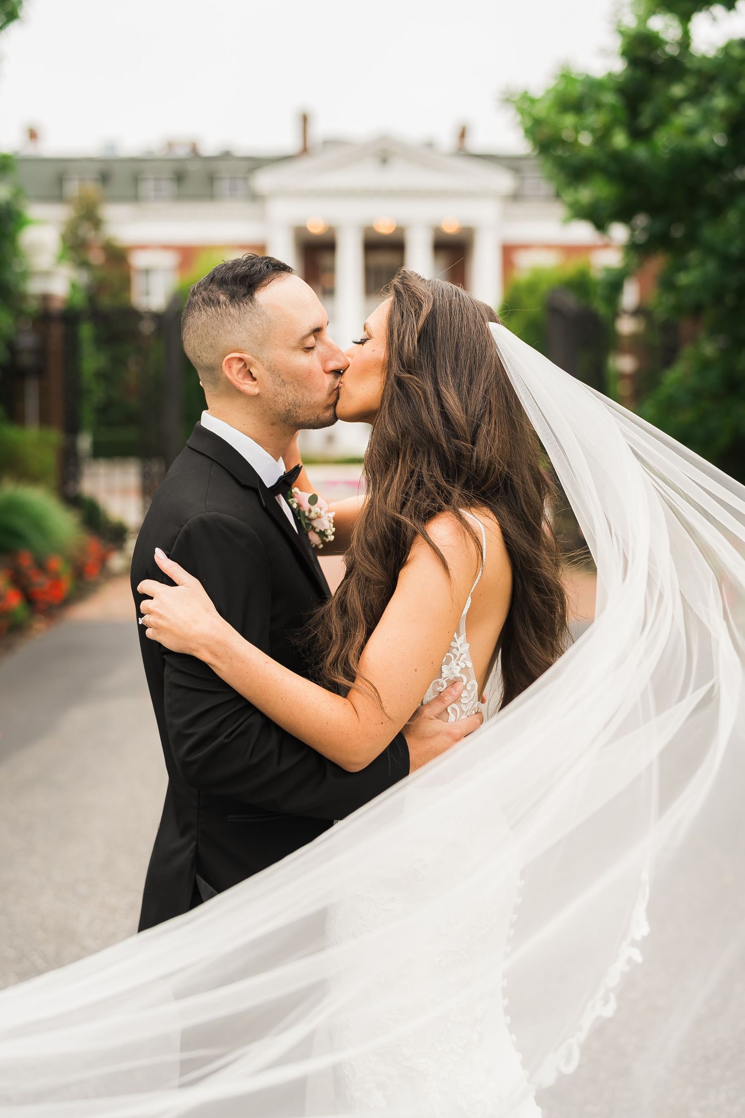 A romantic wedding kiss captured in front of a stately white columned mansion with a flowing veil in the breeze.