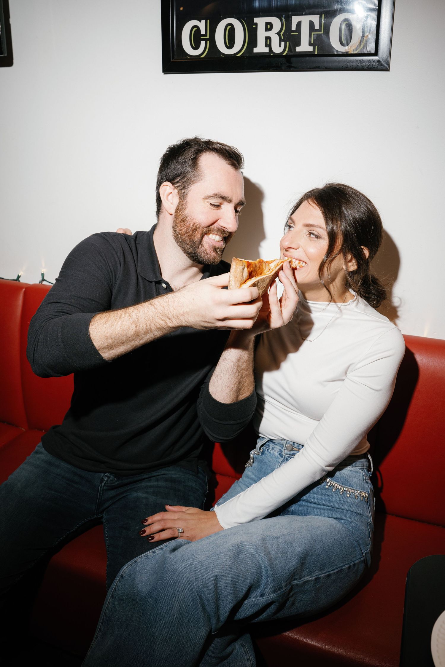 Two people share a laugh while sitting on a red couch under a 'CORTO' sign.