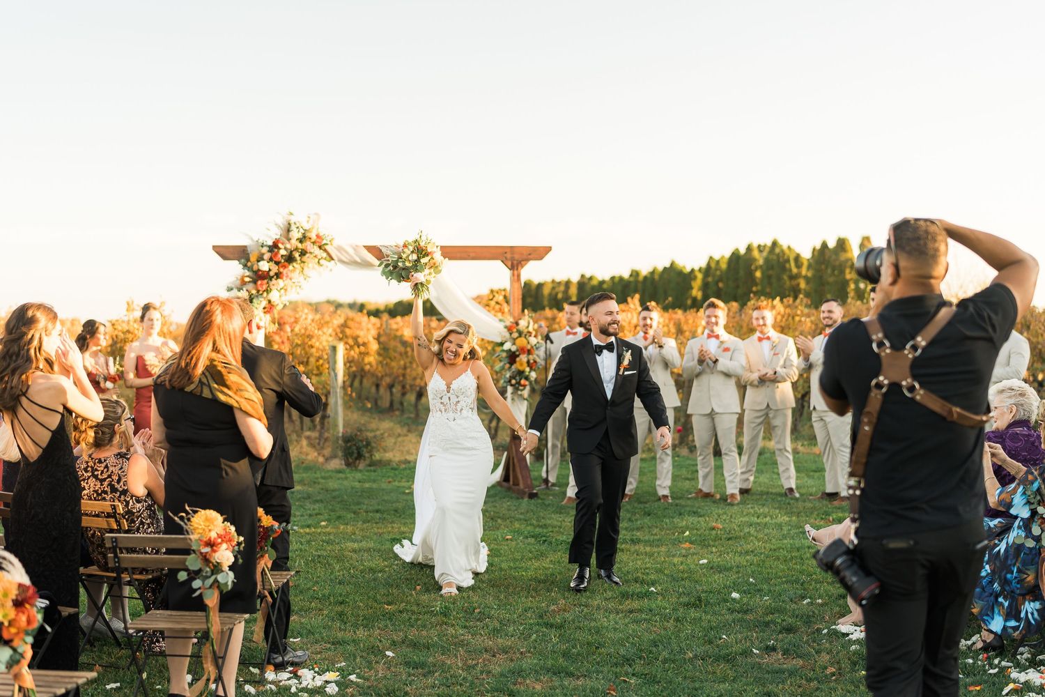 Wedding guests celebrate and throw flower petals as newlyweds walk down outdoor aisle at golden hour ceremony.