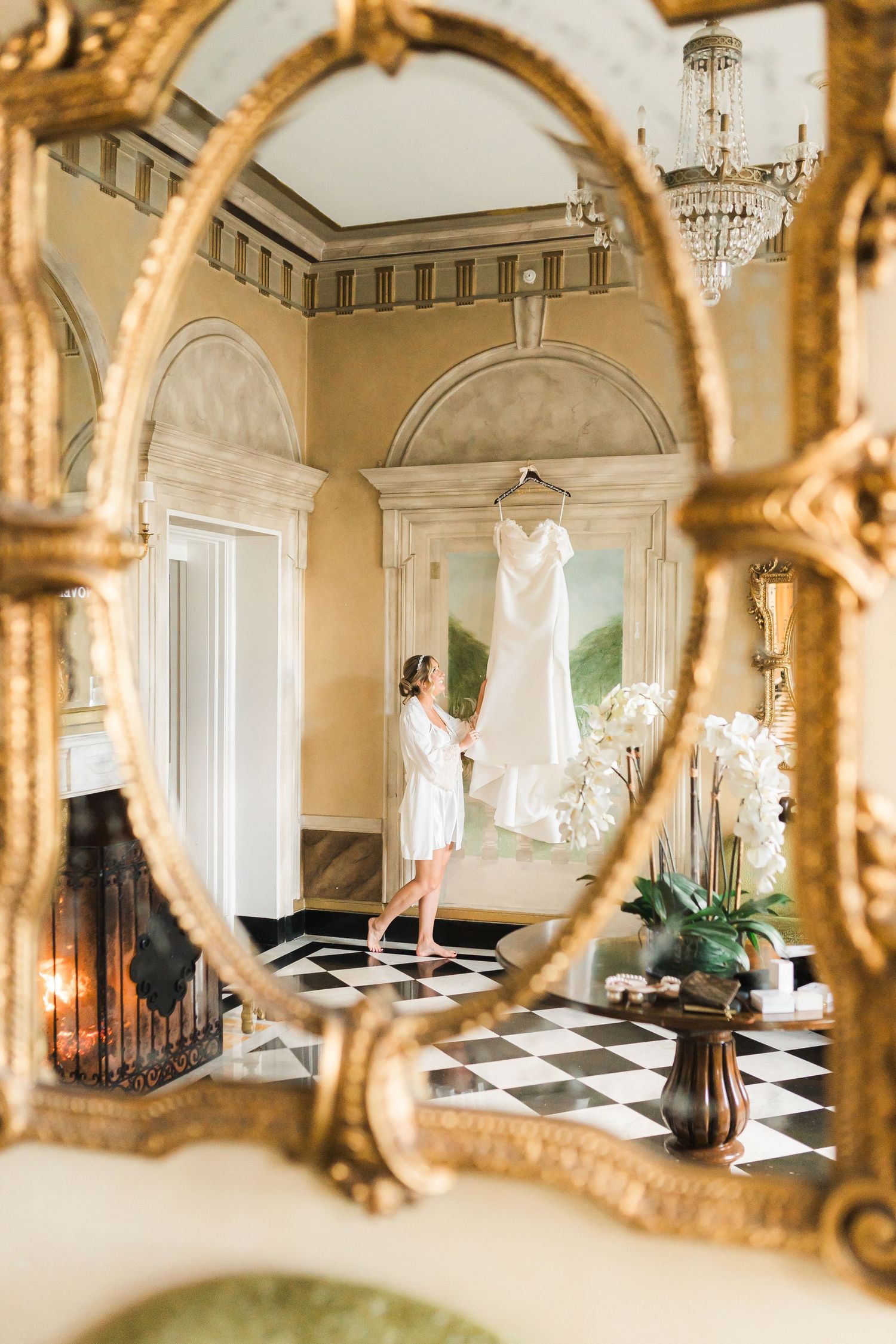 A wedding dress hangs elegantly in an ornate room, reflected in a gold-framed mirror.