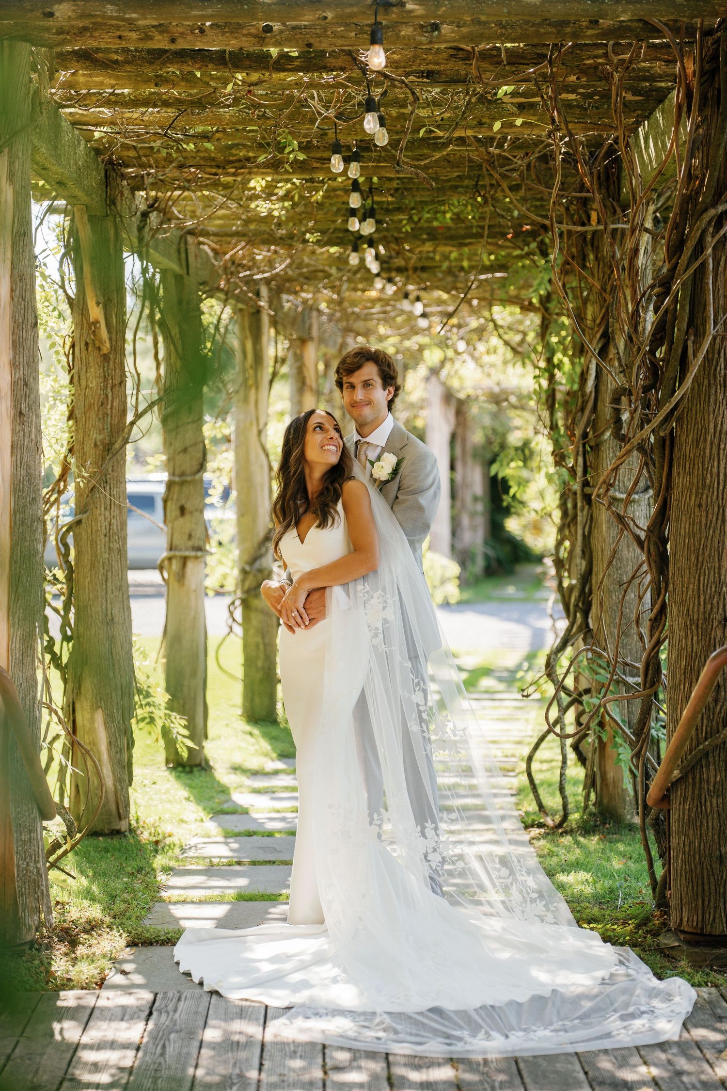 Newlywed couple poses romantically under a vine-covered garden pergola in natural sunlight.