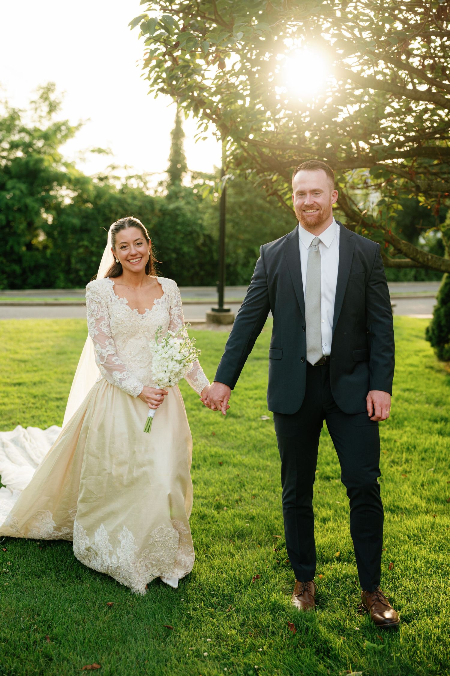A couple poses together on a sunlit lawn in wedding attire during golden hour.