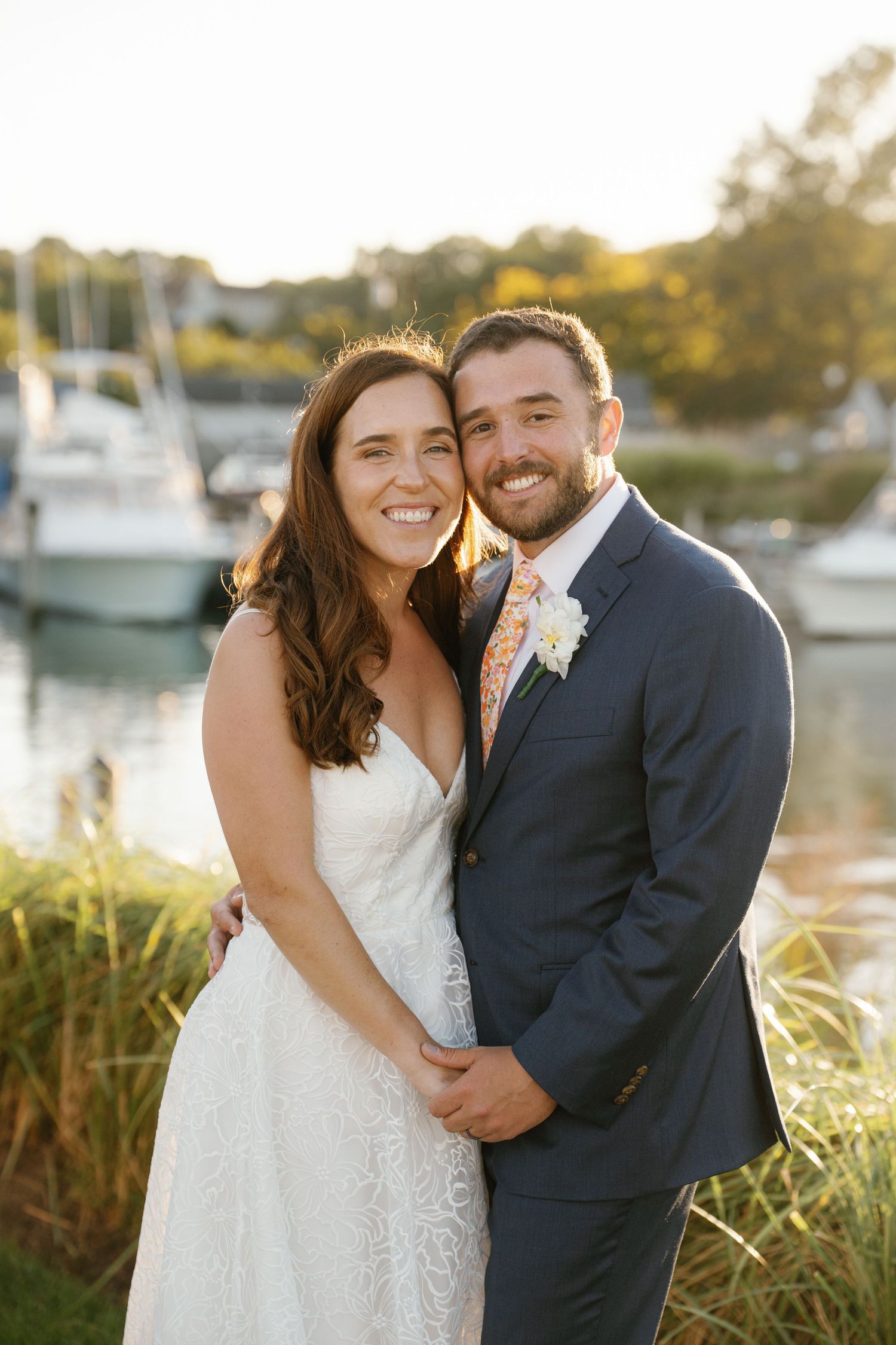 A couple in wedding attire share intimate moments by a marina at sunset with boats in the background.