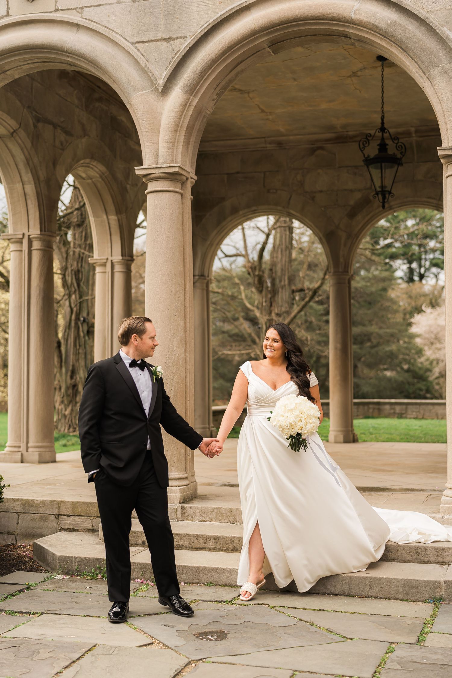 A couple in formal wedding attire hold hands under elegant stone archways with garden views.