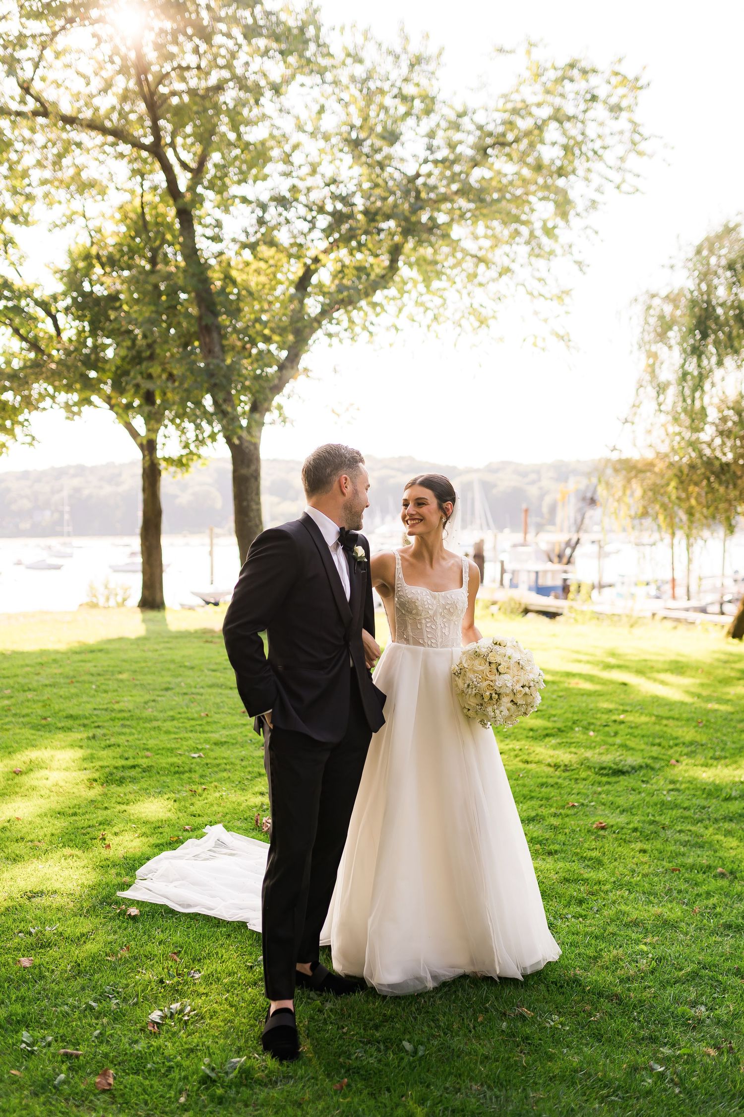 A wedding couple sharing a moment on a sunlit lawn near a waterfront with trees in the background.