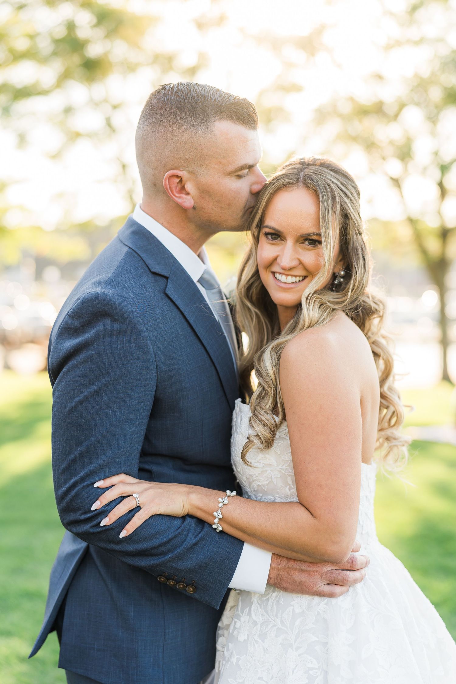 A romantic wedding portrait outdoors shows a couple embracing with the bride in white and groom in navy suit.