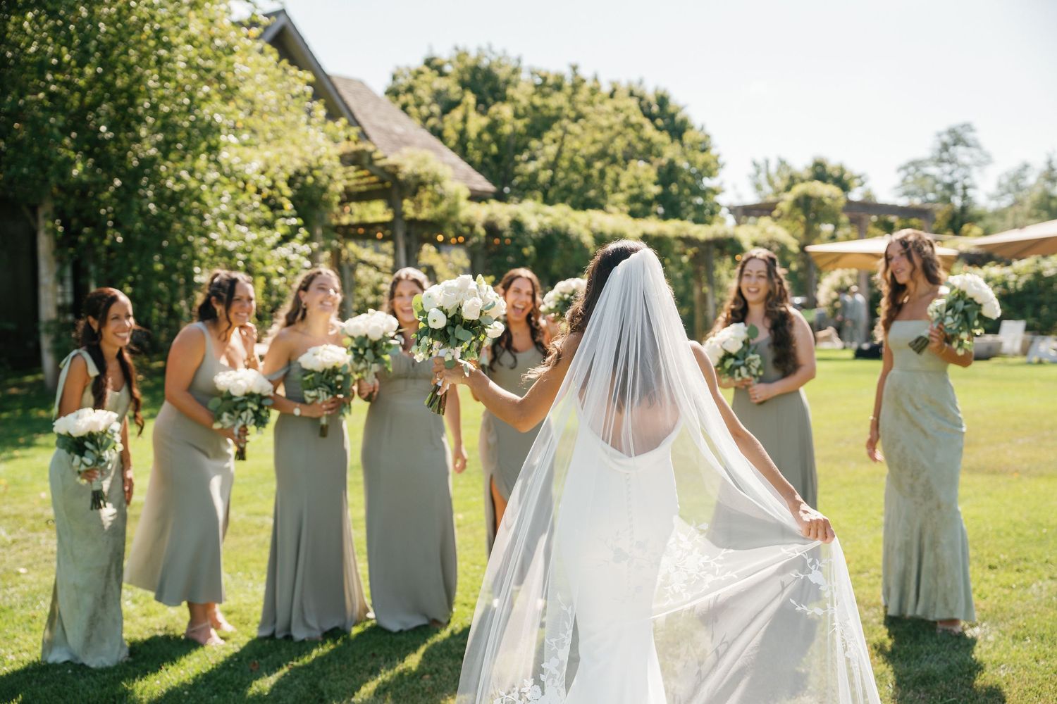 Wedding party in light gray dresses stands together on a sunny lawn during an outdoor celebration.