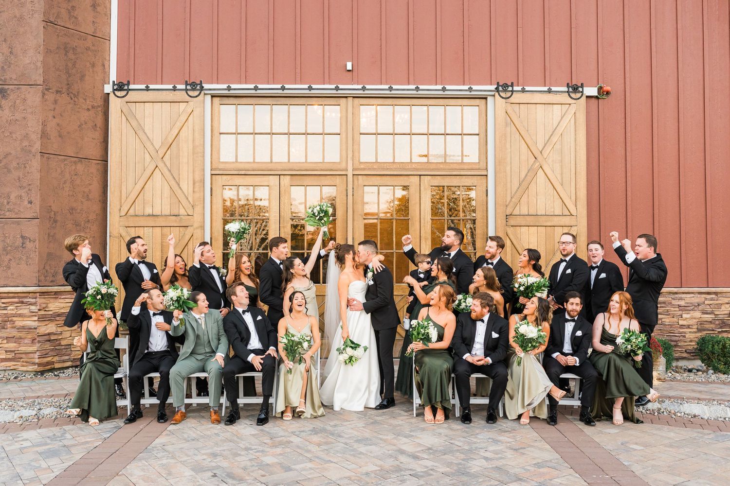 Wedding party in formal attire poses for group photo in front of rustic red barn doors.
