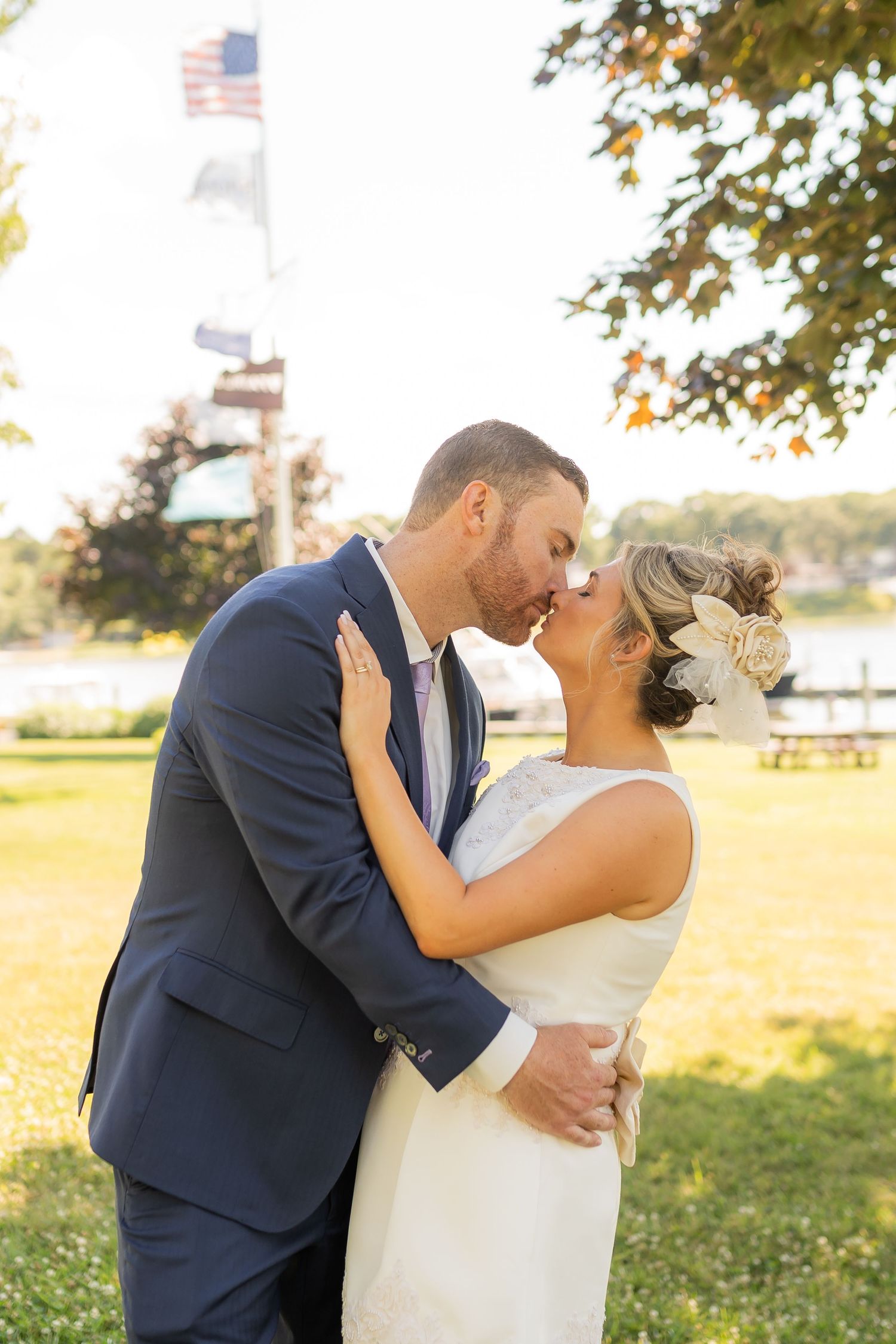 Romantic wedding couple embraces and kisses in a sun-drenched outdoor setting with autumn trees in the background.