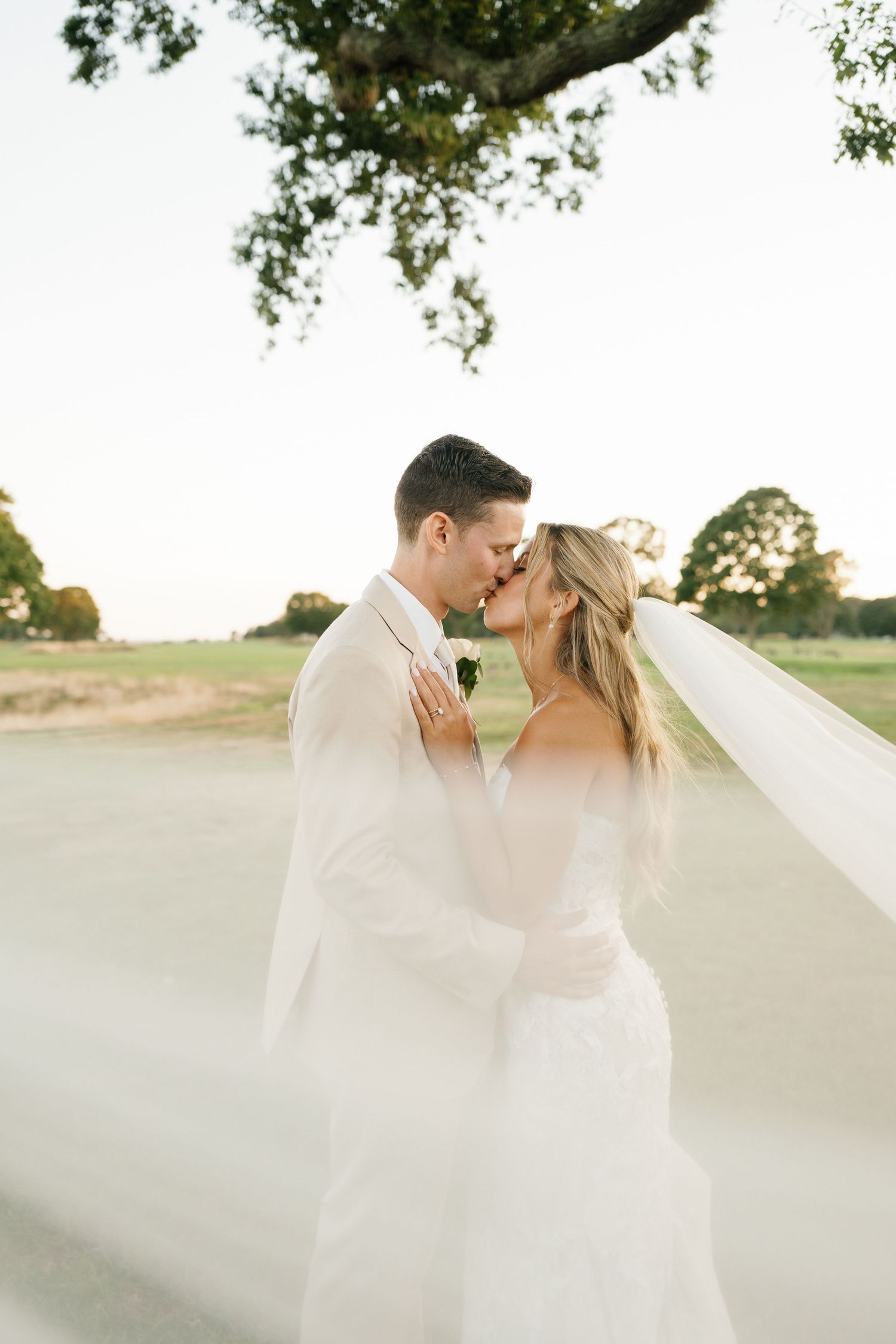 Newlyweds share a romantic kiss outdoors as the bride's veil flows in the breeze at sunset.
