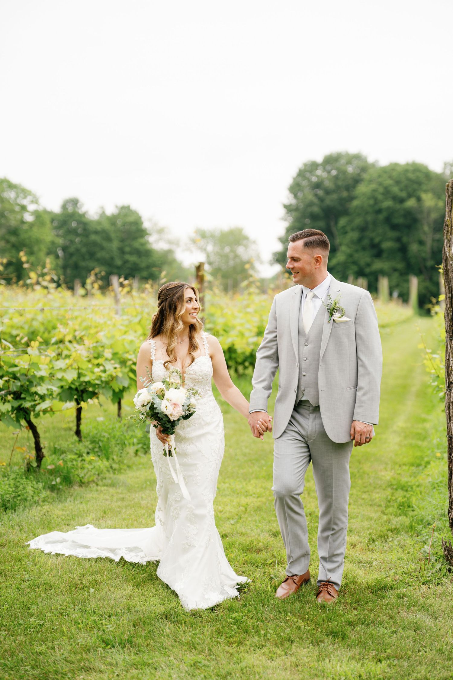 Newlywed couple walks hand in hand through a sunny vineyard on their wedding day in elegant white and gray attire.