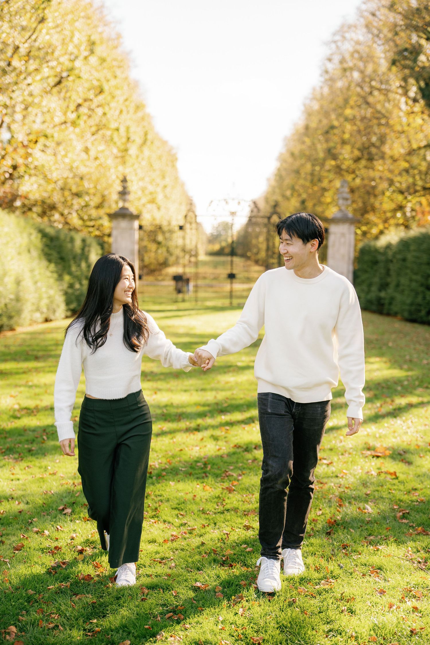 A couple in white and black outfits walk together on a sunny autumn day in a park with golden leaves.