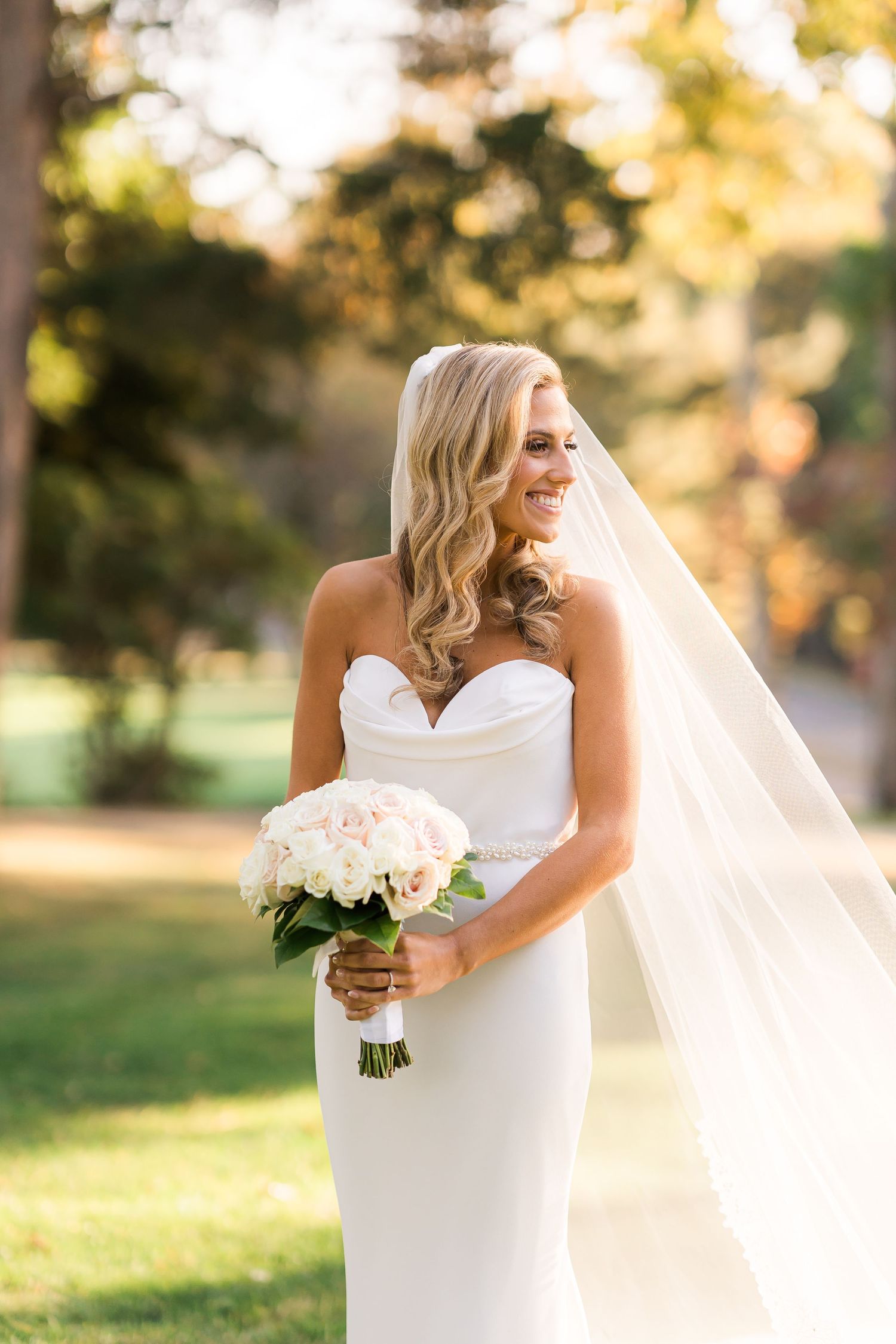 A bride in a fitted white gown holds a classic white bouquet in golden sunlight.