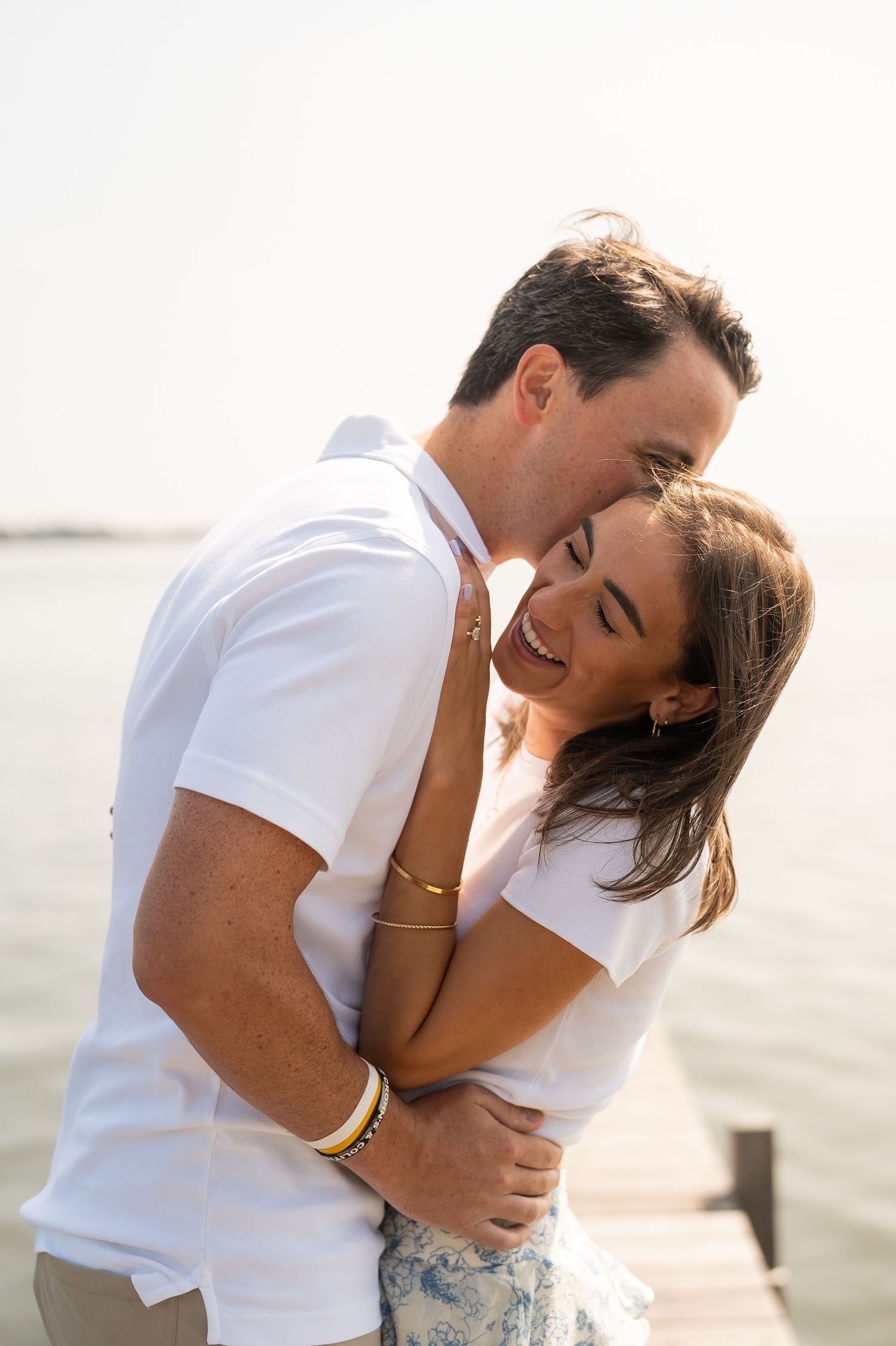 A romantic couple in white shirts embrace and share an intimate moment on a waterfront dock at sunset.