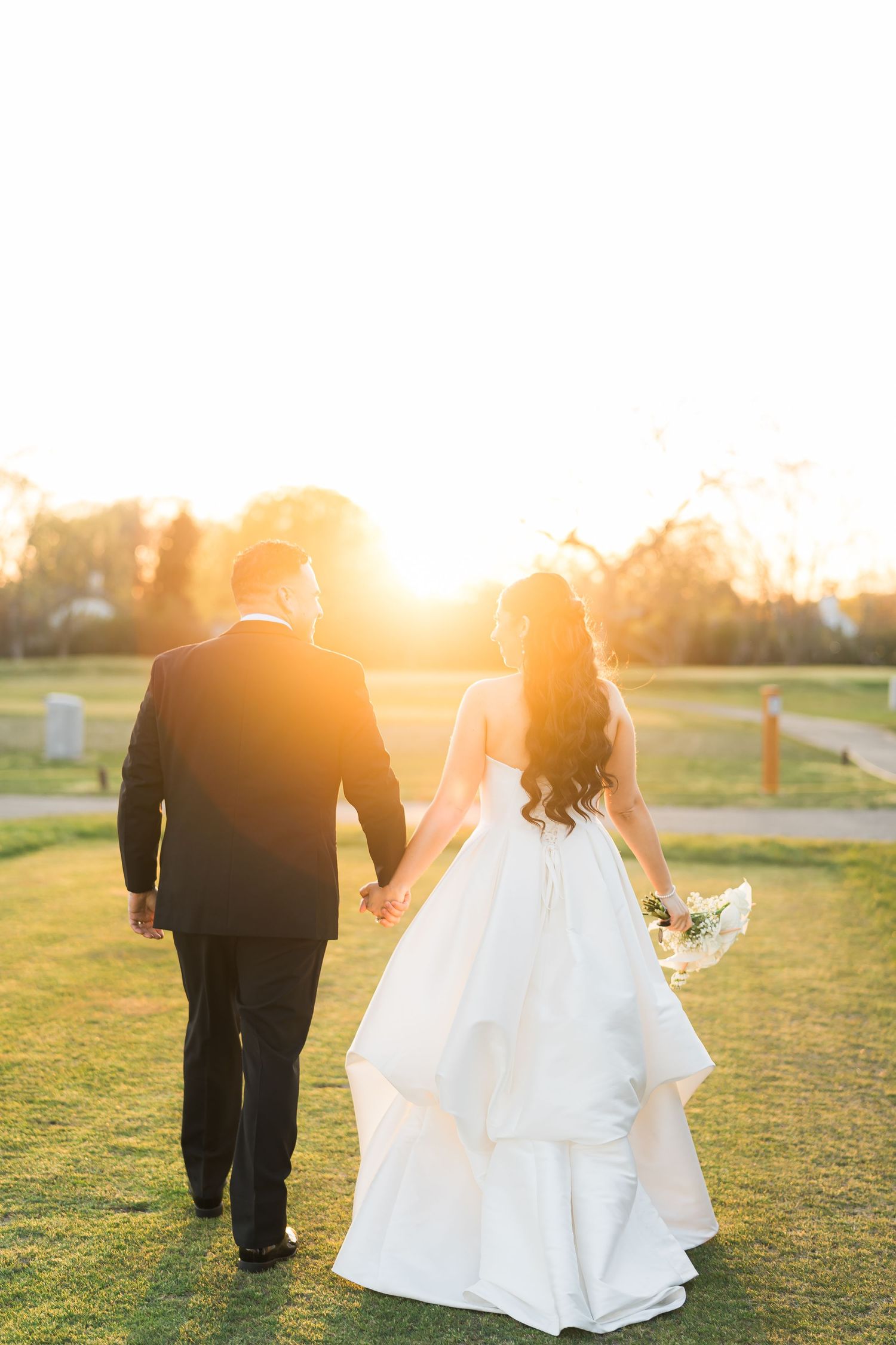 Bride and groom walking hand in hand across a grassy field at sunset with golden sunlight behind them.