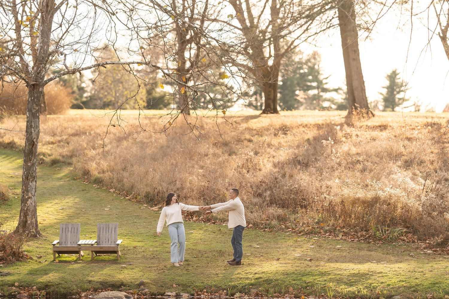 Two people holding hands at sunset in a golden field with Adirondack chairs in the foreground.