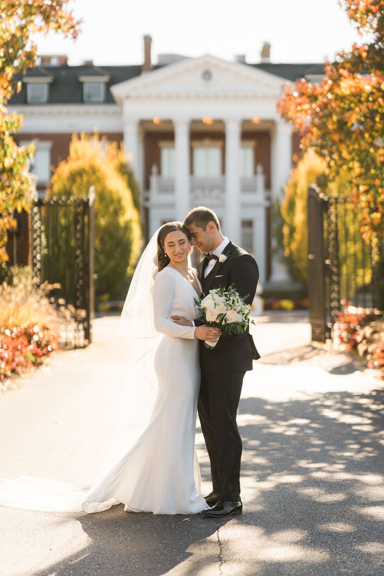 Newlyweds share an intimate moment in front of a grand colonial mansion with autumn foliage.