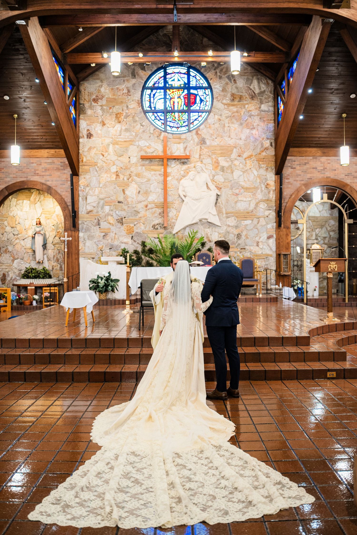 A couple stands at a church altar during their wedding ceremony beneath a stained glass window and wooden beams.