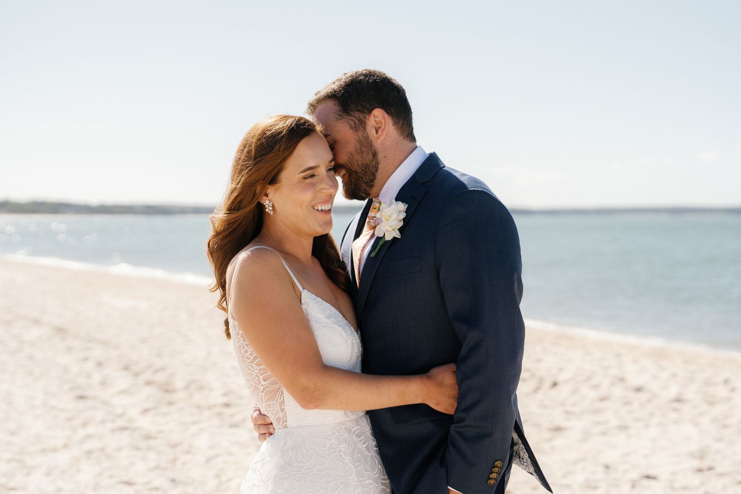 A couple shares an intimate embrace on a sunny beach during their wedding photoshoot.