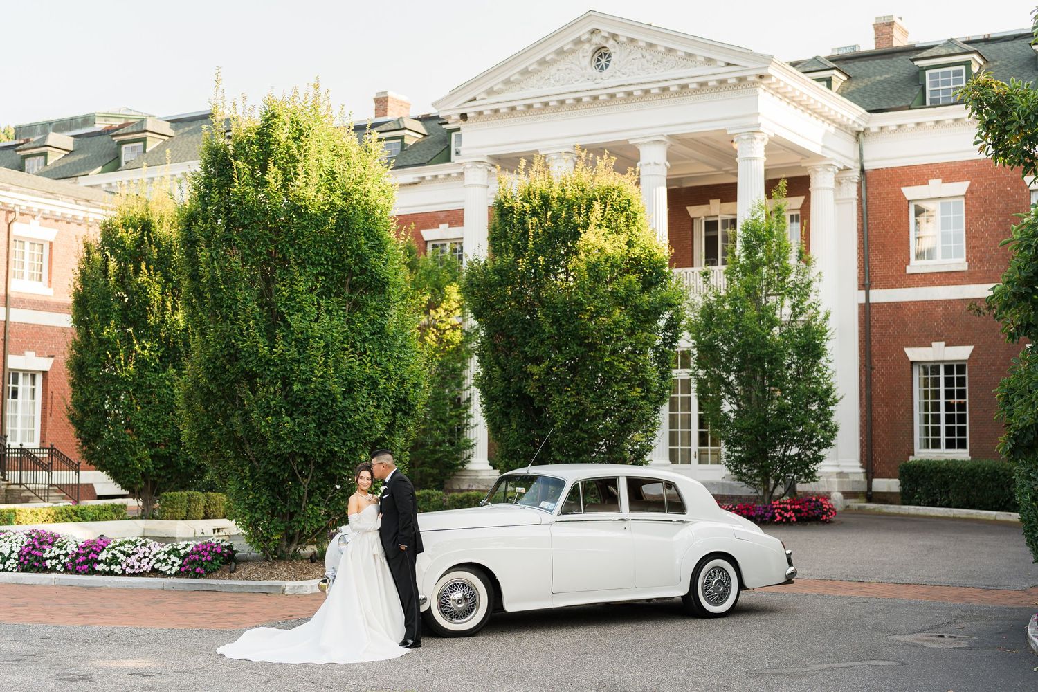 Newlyweds pose beside a classic white vintage car in front of a stately red brick colonial building.