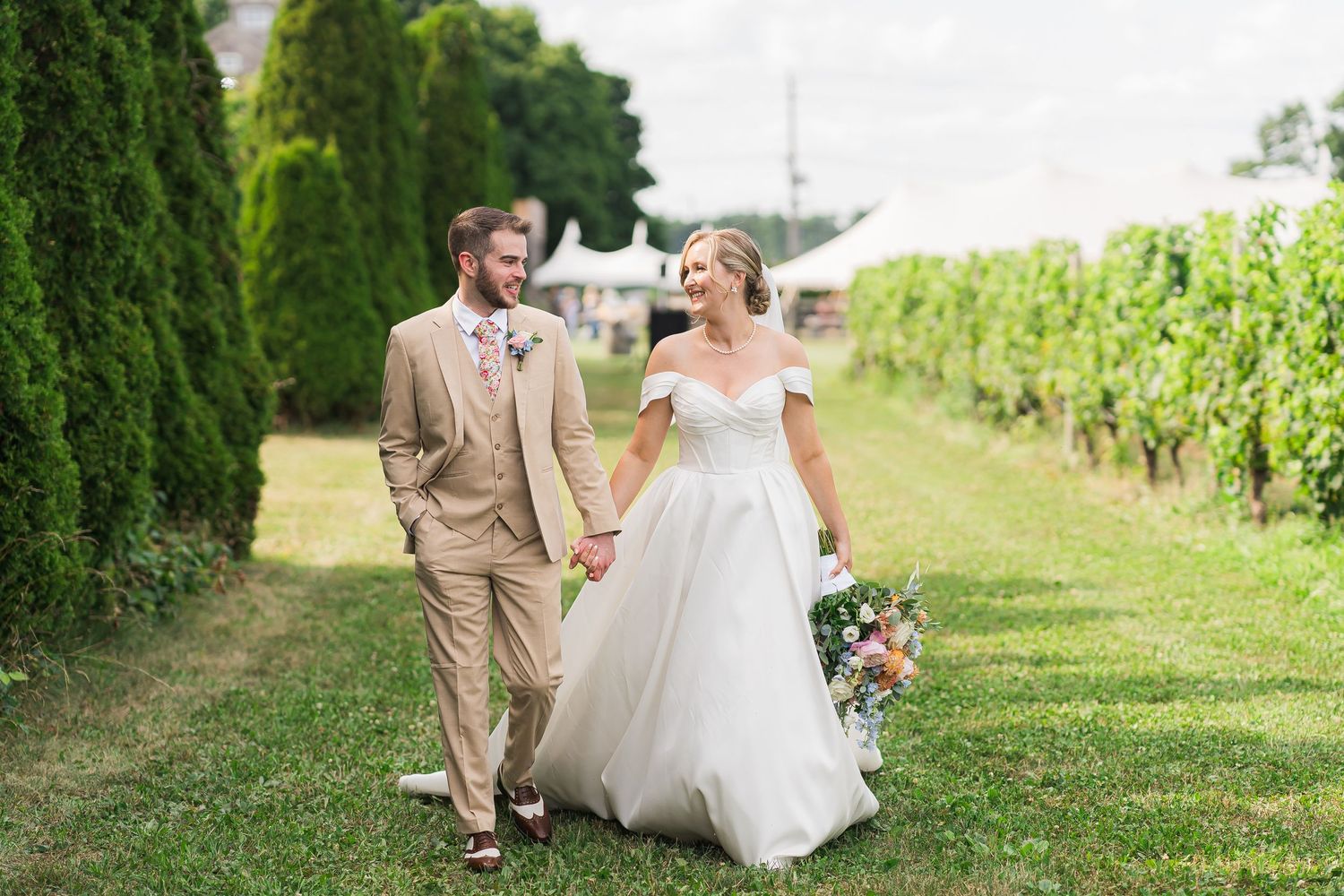 A couple walking together on a grassy path between vineyard rows, celebrating their wedding day in elegant attire.