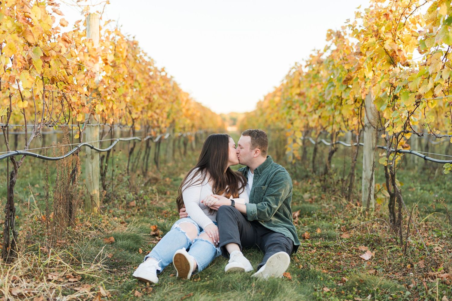 A couple shares a romantic moment while sitting between rows of golden autumn grapevines in a vineyard.