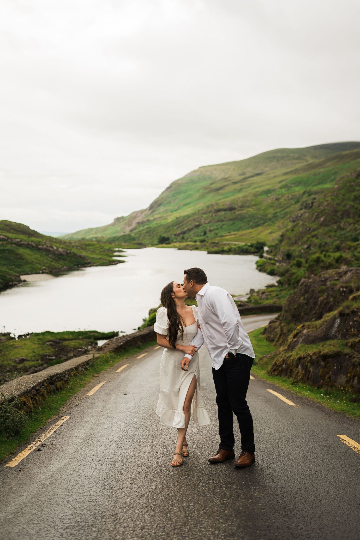 A romantic couple shares a kiss on a winding road alongside a scenic lake in Ireland's lush green countryside.