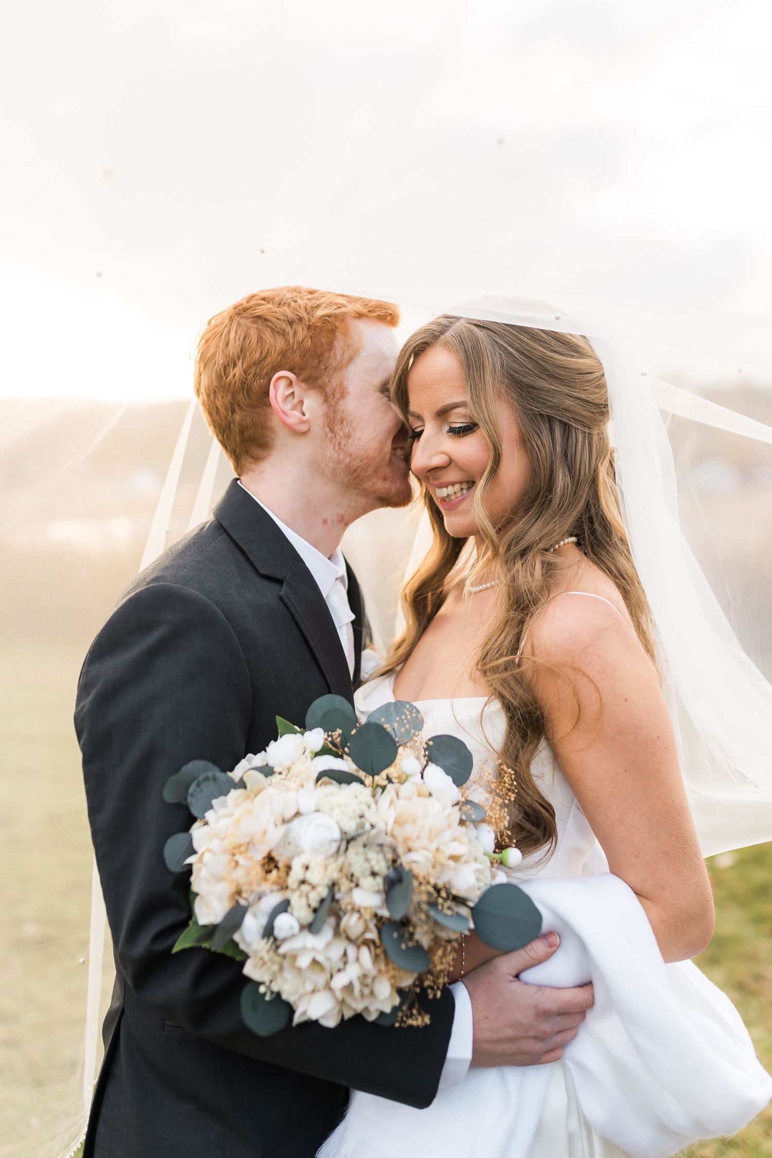 A romantic wedding moment captured outdoors with a beautiful bouquet of white and peach flowers against a soft misty sky.