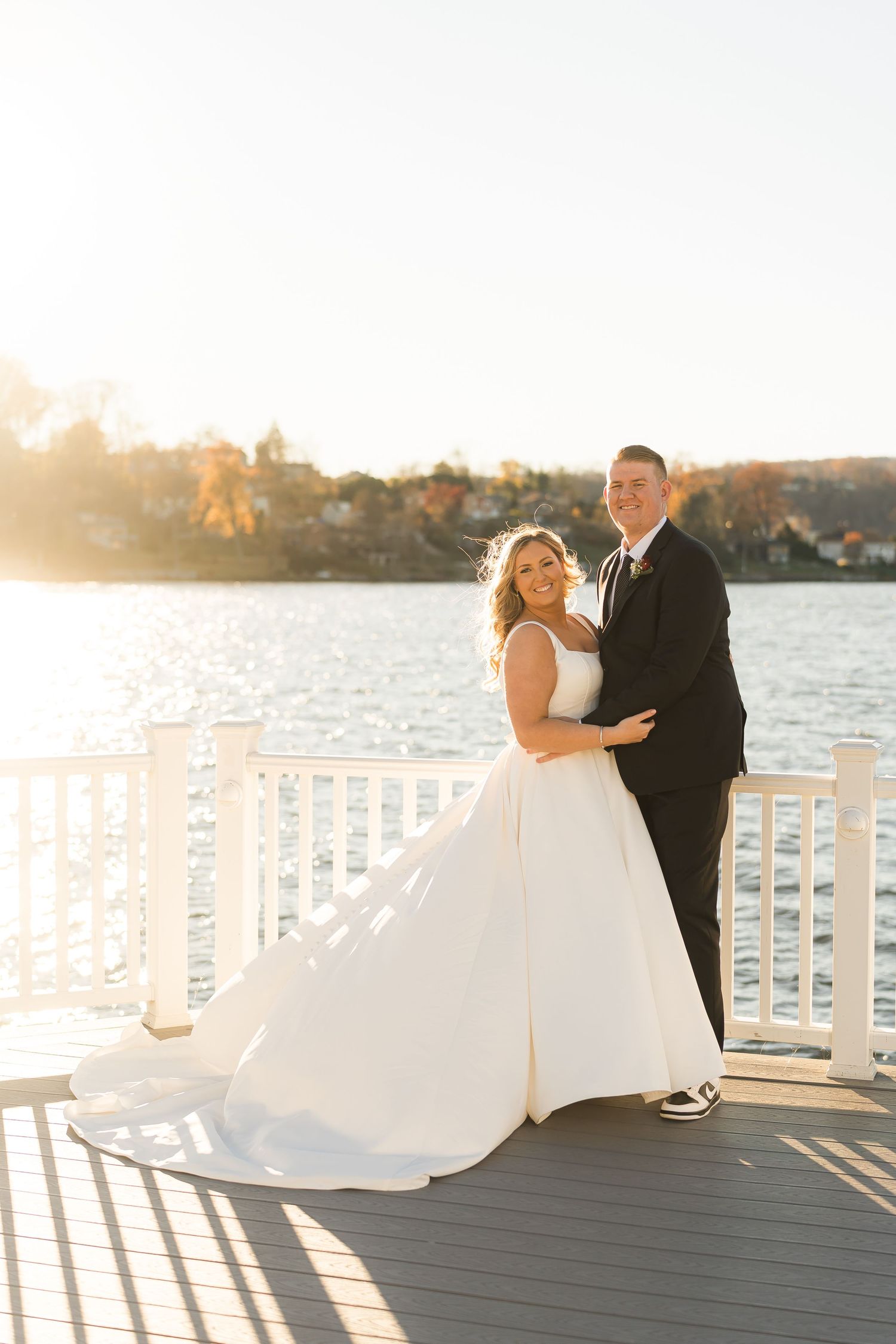 A couple in wedding attire embraces on a waterfront deck during golden hour sunset.