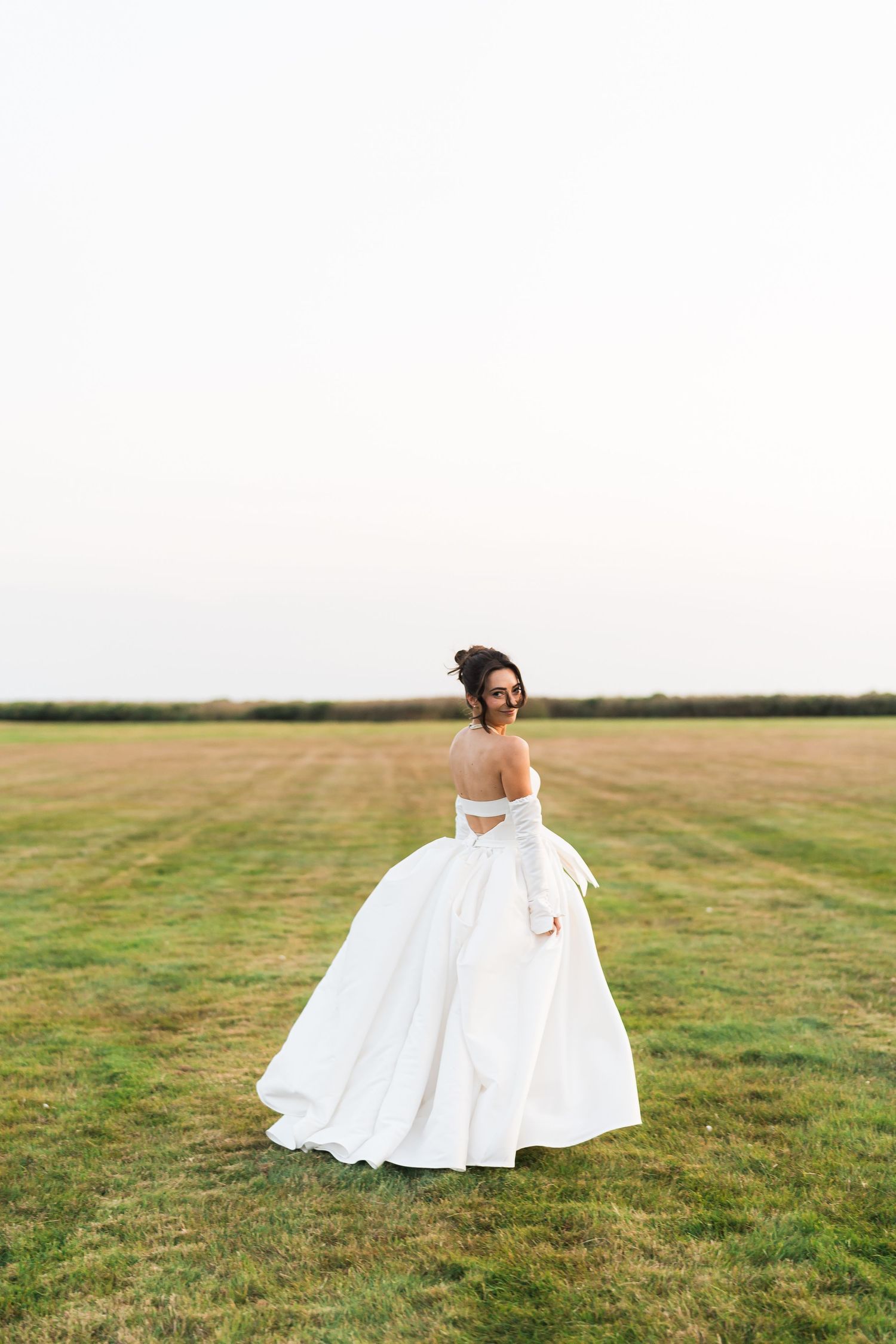 A bride in a flowing white ball gown stands alone in a grassy field on an overcast day.