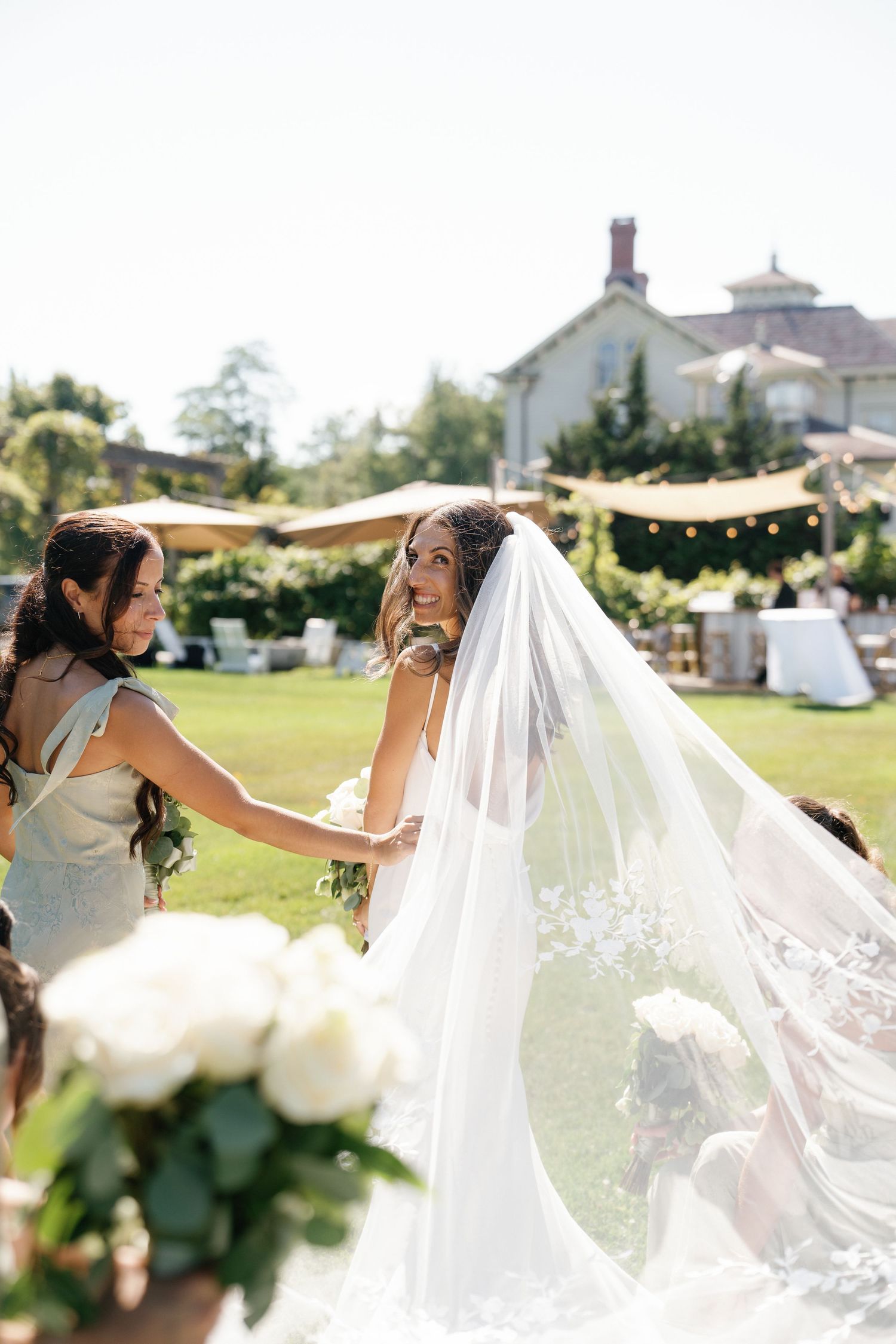 A bride in a flowing white veil twirls joyfully in a sunny garden setting during her wedding photoshoot.