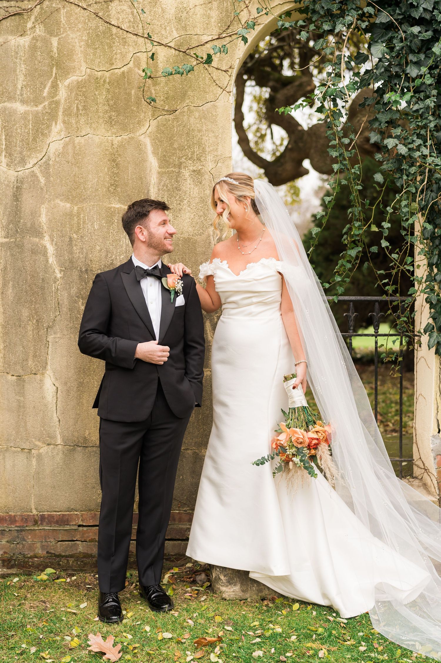 A couple shares a romantic moment against a stone wall with greenery and an archway in an outdoor garden wedding setting.
