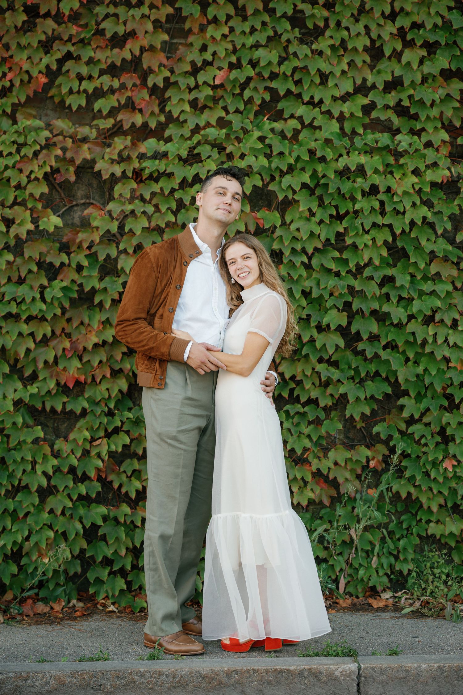 A couple embraces in front of an ivy-covered wall, with the woman wearing a white dress and orange shoes.