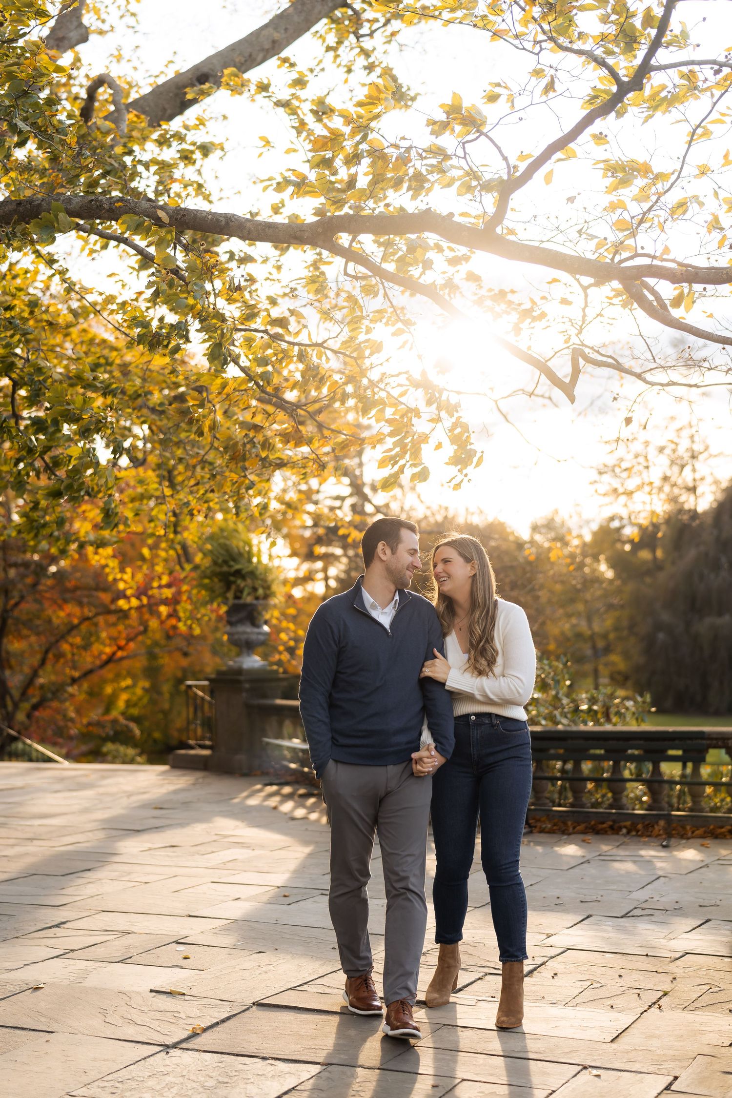 A couple walks arm in arm on a stone path during golden hour with autumn trees and sunlight glowing in the background.