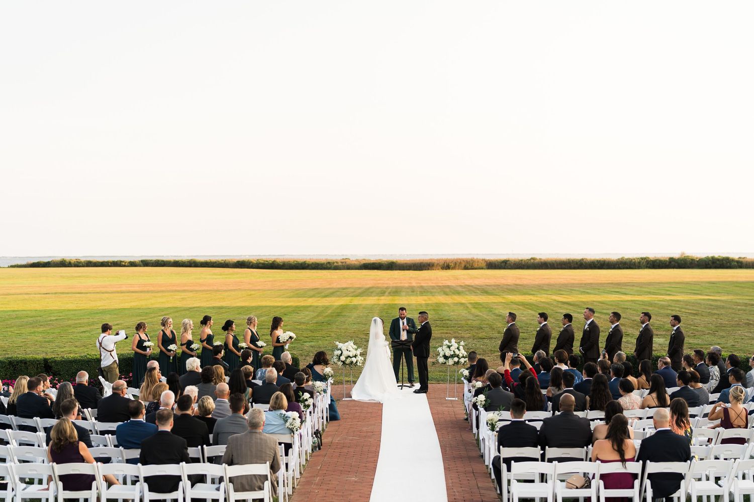 An outdoor wedding ceremony setup with white chairs and an aisle runner against a sweeping field landscape.
